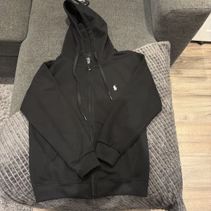 Svart hoodie från Polo Ralph Lauren - Svart hoodie från Polo Ralph Lauren med dragkedja och klassisk vit logga på bröstet. Tröjan har huva med snörning, ribbade muddar och är tillverkad i mjuk bomull. Perfekt för en chill och stilren look.