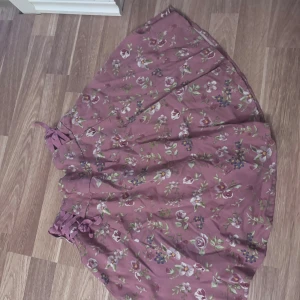 Blommig minikjol från FB Sister - Superfin minikjol i dammigt rosa från FB Sister med blommigt mönster i vitt, grönt och lila. Kjolen har snörning i midjan och ett flowigt fall som ger en romantisk vibe. Perfekt för dig som gillar en feminin och avslappnad stil.