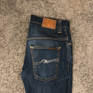 Mörkblå jeans från Nudie Jeans - Snygga mörkblå jeans från Nudie Jeans med klassisk femficksdesign och kontrastsömmar i orange. Bakfickorna har Nudies ikoniska broderade vågmönster. Jeansen är i denim med en rak passform och har läderpatch med logga i midjan. W32 L34