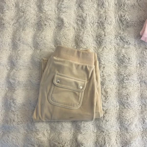 Beiga mjukisbyxor Juicy Couture XS - Beiga mjukisbyxor från Juicy Couture i supermjuk velour med broderad logga på benet. Byxorna har snörning i midjan, bred mudd och ficka med lock och knappar bak. Perfekta för en chill och trendig look.