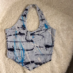 Blå och vit linnetopp från Glitzy  - Säljer en cool linnetopp från Chizzy med abstrakt mönster i blått, vitt och svart. Toppen har ärmlös design och breda axelband, tillverkad i ett syntetmaterial med mesh-liknande struktur. Perfekt för dig som gillar unika och sportiga plagg! Det står M/L men se mått. 