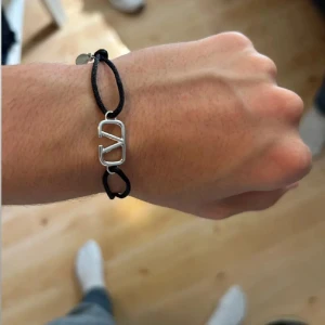 Valentino armband med svart snöre - Snyggt armband från Valentino med svart justerbart snöre och en silverfärgad metallberlock med det ikoniska V-logotypen. Perfekt accessoar för att lyfta din outfit med en stilren och modern touch.