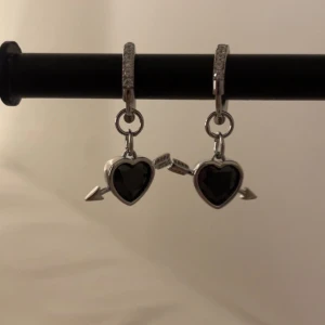 Silverörhängen med svarta hjärtan - Snygga örhängen i silverfärg med svarta hjärtan och pil-detaljer. Ringarna har glittriga stenar och hjärtana hänger som berlocker. Perfekta för dig som gillar unika smycken med lite edge. Rostfri 