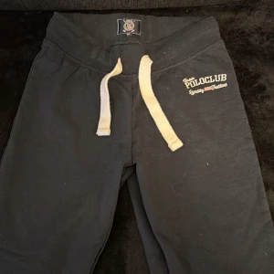 Svarta mjukisbyxor Vinson Polo Club - Svarta mjukisbyxor från Vinson Polo Club med vit snörning i midjan och broderad logga på vänster sida. Byxorna har elastisk midja och är gjorda i mjukt bomullsmaterial, perfekta för chill eller träning.