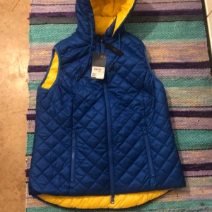 Blå quiltad dunväst från Laurason - Quiltad blå dunväst från Laurason med gul insida och huva. Västen har dragkedja framtill, två fickor med dragkedja och justerbar dragsko vid huvan. Perfekt för lager-på-lager och ger en sportig vibe. Den är storlek M.