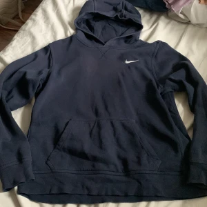 Mörkblå Nike hoodie XL - Snygg mörkblå hoodie från Nike i storlek XL, med klassisk vit Nike-logga broderad på bröstet. Tröjan har huva, magficka och långa ärmar. Perfekt för chill eller sportiga dagar, tillverkad i mjuk bomullsmix för extra komfort. ( XL i barn storlek så sitter som en xs/s) 
