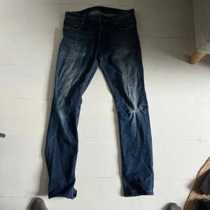 Blå nudie jeans med slitning - Snygga blå nudie jeans med slim passform och slitning på ena benet. Jeansen har en cool tvätt med ljusare partier och är tillverkade i jeansmaterial