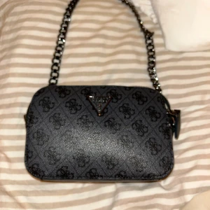 Svart axelväska från Guess med kedja - Snygg svart axelväska från Guess med kedjerem och logomönster över hela väskan. Väskan har två fack med dragkedja och en stilren metallogo framtill. Perfekt storlek för det viktigaste och ger en cool vibe till din outfit.svart/grå