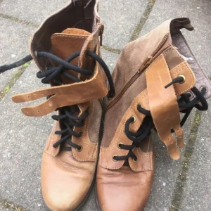 Bruna boots från Dasia - Snygga bruna boots från Dasia med mocca (tror jag att det är men inte 100). De har snörning framtill. Snörena skiljer något i texturen. Dragkedja på sidan och två coola spännen vid skaftet. Rund tå och en liten klack.. Perfekt för dig som gillar detaljer och vill sticka ut. Ingen varm vintersko. En liten nyansskillnad på skorna. Lite slitet skosnöre som fransat sig.