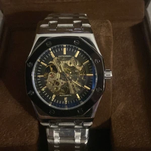 Paterson automatisk klocka silver - Snygg automatisk klocka från Paterson med ett coolt skeletturverk som visar mekaniken. Klockan har en åttakantig boett i silverfärgat stål, svart urtavla med guldiga detaljer och ett robust länkarmband i metall. Perfekt för dig som gillar stil och teknik.