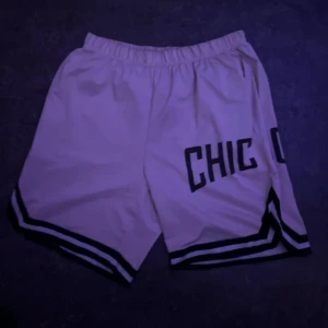 Chicago Bulls basketshorts vita - Säljer ett par vita basketshorts med svart text 'CHICAGO' på sidorna och svarta ränder längs bensluten. De har elastisk midja och är tillverkade i ett lätt polyestermaterial, perfekta för sport eller chill. Klassisk basketstil med loose passform.