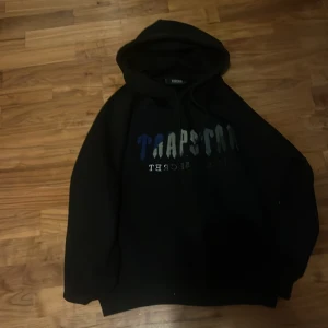 Svart hoodie från Trapstar - Svart hoodie från Trapstar med stor huva och snörning. Framsidan har ett broderat Trapstar-tryck i blått, grått och silver. Mjuk insida och klassisk loose passform, perfekt för streetwear. Snygg detalj med texten 'It's a secret' under loggan.