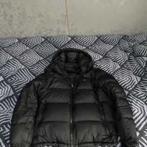 Svart dunjacka från Polo Ralph Lauren - Säljer en svart dunjacka från Polo Ralph Lauren med huva och dragkedja. Jackan har en klassisk pufferstil, två sidofickor med dragkedja och en liten röd logga på bröstet. Nypris 6000kr. Perfekt för kalla dagar och riktigt snygg till streetwear. Köptes förra året. Pris kan sänkas vid snabb affär