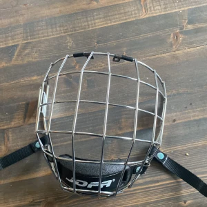 Jofa 480 L hockeygaller - Säljer ett kromat Jofa 480 L hockeygaller i silver metall med svart vaddering för hakan och justerbara remmar. Gallret är CE-märkt och har en robust konstruktion som skyddar ansiktet under hockeyspel. Passar hjälmar med fästen för galler. Intill princip oanvänt och ingen rost!