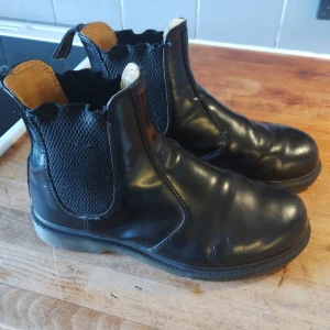 Svarta Dr. Martens chelsea boots stl 37 - Säljer ett par klassiska svarta Dr. Martens chelsea boots i skinn med elastiska paneler på sidorna och dragflikar med gul text bak. Grov sula i gummi och rund tå. Perfekta för dig som vill ha en tidlös och edgy look.