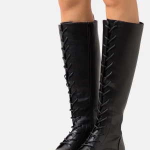 Svarta knähöga boots med snörning - Säljer ett par svarta knähöga boots med grov sula och snörning hela vägen upp. Skorna har rund tå och är gjorda i syntetmaterial med en matt finish. Perfekta för dig som vill ha en edgy look och gillar chunky detaljer.