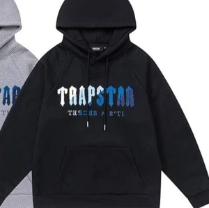 Svart Trapstar hoodie med blå text - Säljer en svart hoodie från Trapstar med fet logga i vitt och blått framtill. Hoodien har huva med snörning och en stor magficka. Perfekt för dig som vill ha en streetstyle-look med edge.