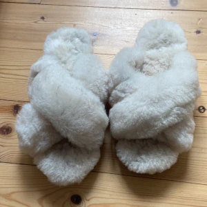 Fårskinnstofflor från Canada Snow - Supermysiga och fluffiga tofflor från Canada Snow i ljusbeige fårskinn. De har korslagda band över foten och en platt sula med bra grepp. Perfekta för att hålla fötterna varma hemma under kyliga dagar.