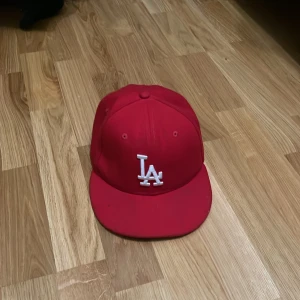 Röd LA-keps från New Era 59FIFTY - Snygg röd keps från New Era 59FIFTY med LA-logga i vitt framtill och MLB-logga bak. Klassisk rak skärm och broderade ventilationshål. Perfekt för dig som vill ha streetstyle med sportig vibe.