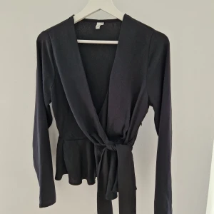 Svart omlottblus från NLY Trend - Svart omlottblus från NLY Trend i storlek 34. Blusen har långa ärmar, knytband i midjan och lätt volang nertill. Tillverkad i mjuk polyester med djup v-ringning och stilren look.