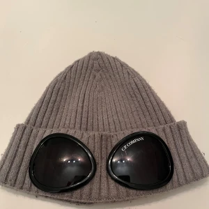 Grå ribbad Goggle-mössa från C.P. Company - Bara använt 3 gånger. Säljer en grå ribbstickad Goggle-mössa från C.P. Company med svarta glasögon-detaljer framtill. Mössan har en uppvikt kant och är tillverkad i mjukt material som håller dig varm och ger en unik streetwear-look.