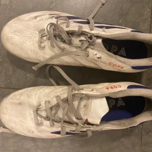 Adidas Copa vita fotbollsskor - Adidas Copa fotbollsskor i vitt med blå och röda detaljer. Skorna har snörning och en platt sula med fasta dobbar i svart och rött. Ovandelen är i skinn och insidan har blå textil med Copa-tryck. Perfekt för dig som vill ha klassiska och snygga fotbollsskor.