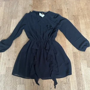 Svart volangklänning från Kappahl - Supersnygg svart kortklänning från Kappahl med långa ärmar och volangdetaljer framtill. Klänningen har omlottliknande v-ringning och resår i midjan för en skön passform. Materialet är lätt och luftigt, perfekt för dig som gillar stilrena vibes.