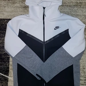 Nike Tech fleece  - Nike Tech fleece med dragkedja i vitt, svart och grått. Snyggt V-format färgblocksmönster, huva och lång ärm. Svart Nike-logga på bröstet och praktisk ficka med dragkedja på ärmen. Perfekt för en sportig och chill stil.