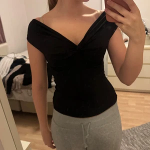 Svart omlott topp med offshoulder - Säljer en svart omlott topp med snygg offshoulder-design och v-ringad fram. Toppen är tight och sitter figurnära, perfekt för dig som gillar en stilren och enkel look. Materialet känns mjukt och stretchigt, vilket gör den bekväm att bära. Passar dig som vill ha en klassisk svart topp med lite edge. Använd en gång
