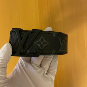 Svart bälte med monogram - Svart bälte med klassiskt -monogram och ett -spänne i mörk metall. Bältet har en bred och stilren form, med diskret mönster i grått och svart. Perfekt för dig som vill ha en lyxig och ikonisk accessoar.