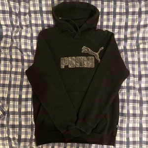 Svart hoodie från Puma med logga - Svart hoodie från Puma med stor logga och puma-tryck på bröstet. Tröjan har huva med snörning och ribbade muddar vid ärmslut och nederkant. Materialet är mjukt och känns skönt mot huden, perfekt för chill eller streetwear. Snyggt mönstrad logga i grått.