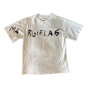 Vit Maison Margiela MM6 t-shirt - Vit t-shirt från Maison Margiela MM6 med svart grafiskt tryck över bröstet. Klassisk passform med rund hals och korta ärmar. Tillverkad i mjuk bomull och har en edgy, handskriven logga framtill. Perfekt för dig som gillar statement-plagg.