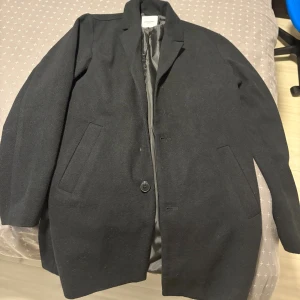 Svart rock från Jack & Jones, S - Stilren svart rock från Jack & Jones i storlek S. Klassisk design med krage, två sidofickor och knappstängning framtill. Yttermaterialet har en ullig känsla och insidan är fodrad med slätt tyg. 