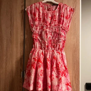  rosa blommig kortklänning - Supersöt kortklänning från BikBok i rosa och rött med blommigt mönster. Klänningen har volangdetaljer. Den är ärmlös och har en rund halsringning. En helt perfekt klänning för sommaren! Aldrig använd, lappen kvar!