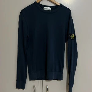 Mörkblå Stone Island sweatshirt - Säljer en mörkblå sweatshirt från Stone Island med den klassiska loggan på vänster ärm. Tröjan har rund halsringning, långa ärmar och ribbade muddar. Perfekt för dig som gillar stilrena och ikoniska plagg.