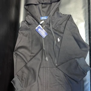 Svart hoodie  - Svart hoodie med dragkedja framtill, huva med snörning och klassisk logga broderad på bröstet. Tröjan har ribbade muddar och känguruficka. Perfekt för en chill och stilren look.