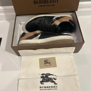 Burberry sneakers med klassiskt rutmönster - Snygga sneakers från Burberry med svart skinn och det ikoniska beigea rutmönstret på sidorna. Skorna har svarta snören, vit sula och Burberry-logga bak på hälen. Perfekta för dig som vill ha en stilren och lyxig look.