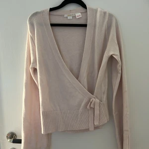 Ljusbeige omlottkofta från Gina Tricot - Supersöt ljusbeige omlottkofta från Gina Tricot med knyt i sidan och bred ribbad mudd nedtill. Koftan har lång ärm och är stickad i mjukt material, perfekt för lager på lager under kyliga dagar.