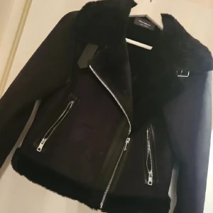 Svart pälsjacka från Pull&Bear - Säljer en svart pälsjacka från Pull&Bear med stora slag, fuskpälsdetaljer och silvriga dragkedjor. Jackan har två snedställda fickor med dragkedja och en snygg spänndetalj vid kragen. Perfekt för kalla dagar och riktigt snygg till jeans.