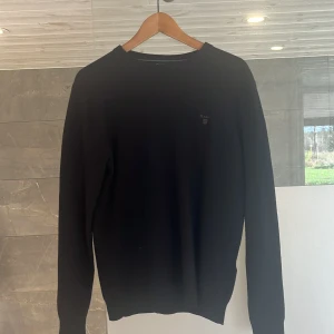 Svart ulltröja från GANT, storlek L - Klassisk svart tröja från GANT i mjuk lammull. Enkel design med rund halsringning och diskret logga på bröstet. Perfekt för lager-på-lager och funkar året runt. Långärmad och ribbade muddar för en snygg passform. Passar M mycket bra också. 