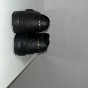 Svarta Alexander McQueen sneakers - Säljer ett par svarta sneakers från Alexander McQueen med guldfärgad logga på hälen. Skorna har en chunky plattformssula, rund tå och snörning framtill. Tillverkade i skinn med stilren design och diskreta sömmar. Perfekta för dig som gillar exklusiva och trendiga sneakers.