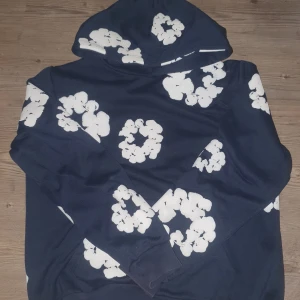 Mörkblå hoodie från Denim Tears - Säljer en mörkblå hoodie från Denim Tears med stora vita blommönster över hela tröjan. Tröjan har huva och långa ärmar, tillverkad i mjuk bomull. Perfekt för dig som vill sticka ut med en unik och trendig look.