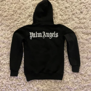Svart Palm Angels hoodie med tryck - Svart hoodie från Palm Angels med stor vit logga på ryggen och mindre logga på bröstet. Klassisk känguruficka framtill, snörning i huvan och ribbade muddar. Perfekt för dig som gillar streetwear och vill sticka ut med en ikonisk look storleken är s i barn och är 60cm lång och 43cm bred
