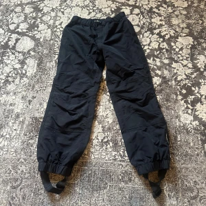 Svarta täckbyxor från Kaxs Proxtec - Svarta täckbyxor från Kaxs Proxtec i storlek 128. Byxorna har resår i midjan och vid bensluten, samt praktiska fothällor och reflexdetaljer på benen. Tillverkade i slitstarkt syntetmaterial som står emot väta och vind, perfekta för kalla dagar.