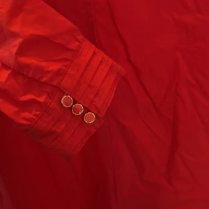 Röd långärmad klänning från Culture -  ALDRIG ANVÄND!!! Ny pris 1100kr.  Röd klänning från Culture med lång ärm, v-ringning och snygga guldknappar vid ärmslutet. Klänningen har en rak passform och är tillverkad i ett slätt bomullsliknande material. Perfekt för dig som vill sticka ut med färg och stil.