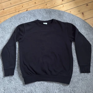Svart sweatshirt från KappAhl i storlek 158/164 (XXS) - En stilren svart sweatshirt från KappAhl i mjuk bomull. Tröjan har rund halsringning, ribbade muddar vid ärmslut och nederkant samt lång ärm. Perfekt för en clean och enkel look. Anledningen till att jag säljer den, är pga utväxt. Annars fin skick. 