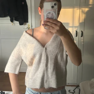 Beige ribbad stickad blus med knappar - Supermysig beige stickad blus med ribbat material och knappar framtill. Modellen är croppad med v-ringning och trekvartsärm, perfekt för lager-på-lager och chill vibes. Snygg att bära off-shoulder för en avslappnad look.