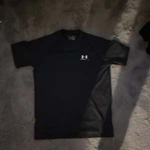 Svart Under Armour t-shirt i polyester - Svart t-shirt från Under Armour med vit logga på bröstet och i nacken. Tillverkad i mjuk och lätt polyester som andas, perfekt för träning. Klassisk rund hals och korta ärmar, med en slim passform som sitter snyggt på kroppen. Lite sönder men den funkar perfekt.