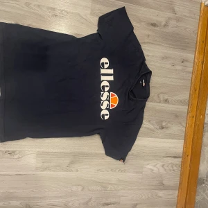 Svart t-shirt från Ellesse, storlek L - Snygg svart t-shirt från Ellesse i storlek L. T-shirten har ett stort Ellesse-tryck i vitt och orange på bröstet. Klassisk rund hals och korta ärmar. Tillverkad i 100% bomull för skön och mjuk känsla. Perfekt för dig som gillar sportiga och stilrena plagg.