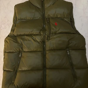 Olivgrön Polo Ralph Lauren väst  - olivgrön väst, Västen har hög krage, två sidofickor med dragkedja. Perfekt att slänga över en hoodie för extra värme. Västen är för liten på mig men passar dig som har M i storlek 